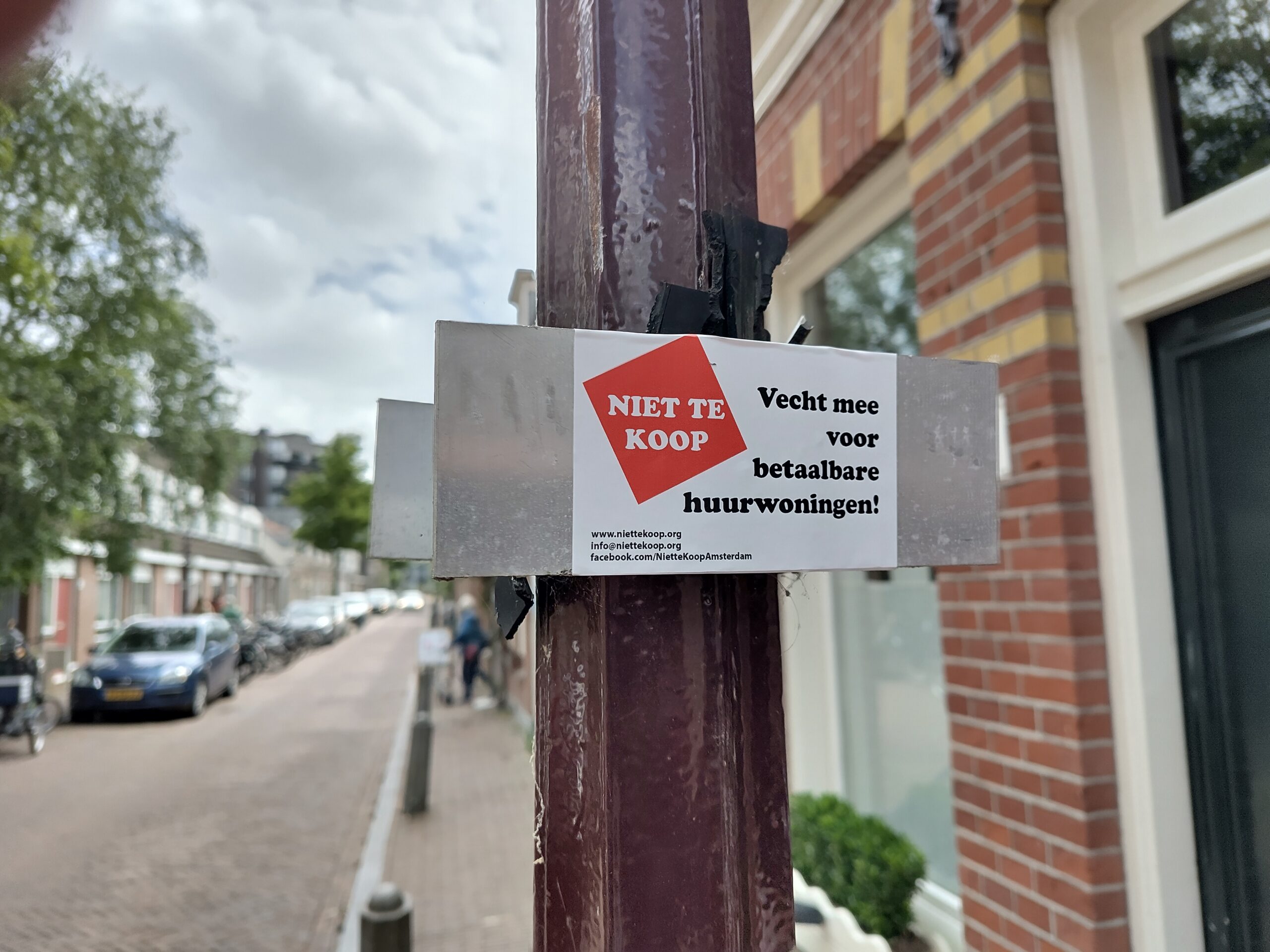 Sticker van actiegroep #NietteKoop op een bordje op een paal. In de achtergrond de straat te zien.
