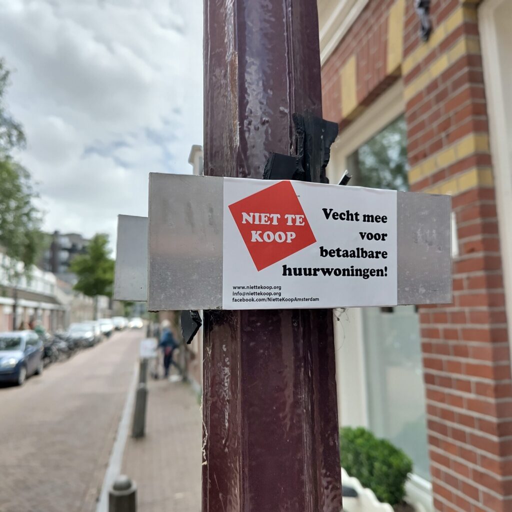 Sticker van actiegroep #NietteKoop op een bordje op een paal. In de achtergrond de straat te zien.